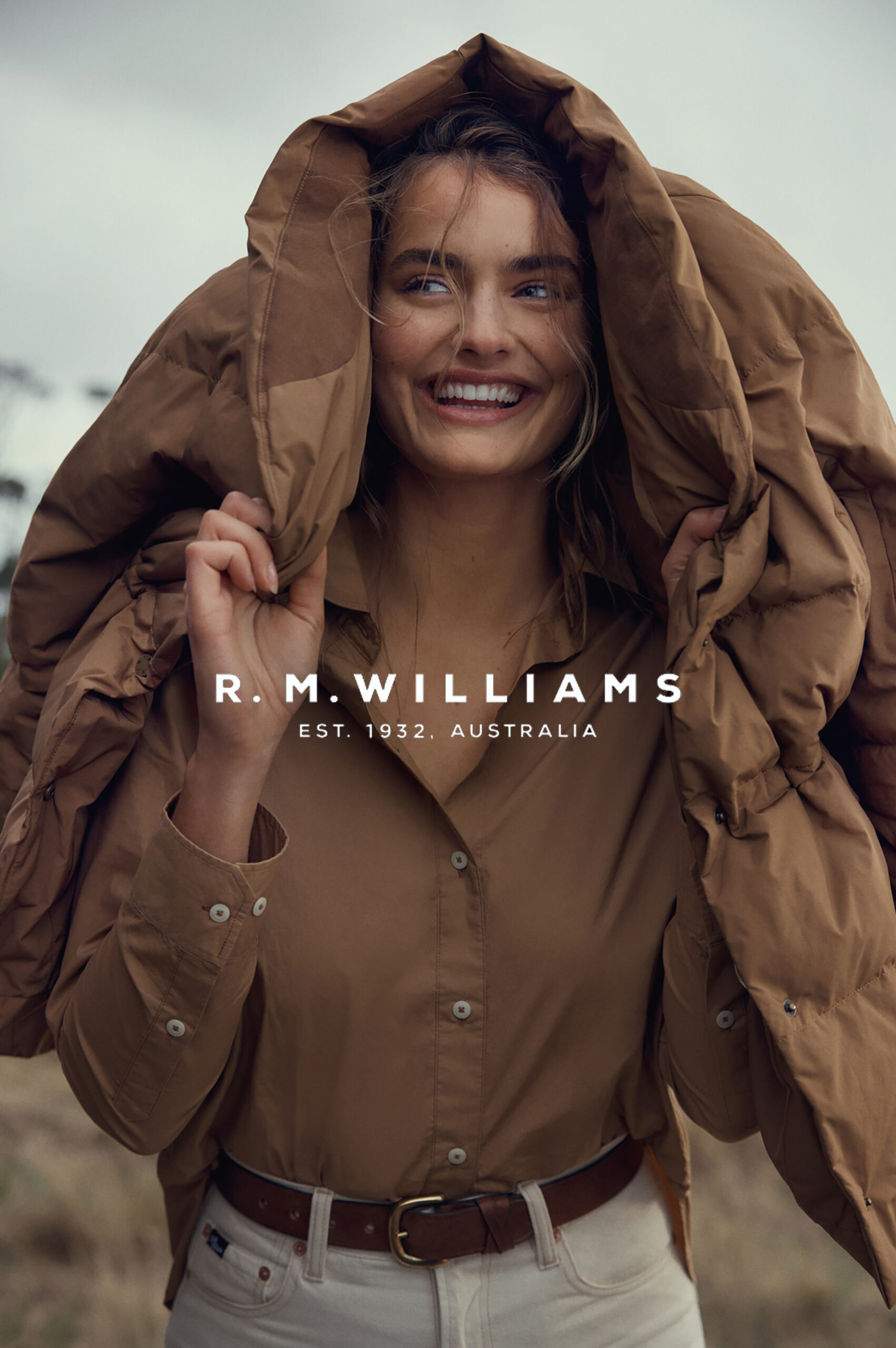 Smiling adult female in R.M. Williams outdoor fashion ad. She wears a brown button-down shirt, white pants, and a puffy jacket hood.