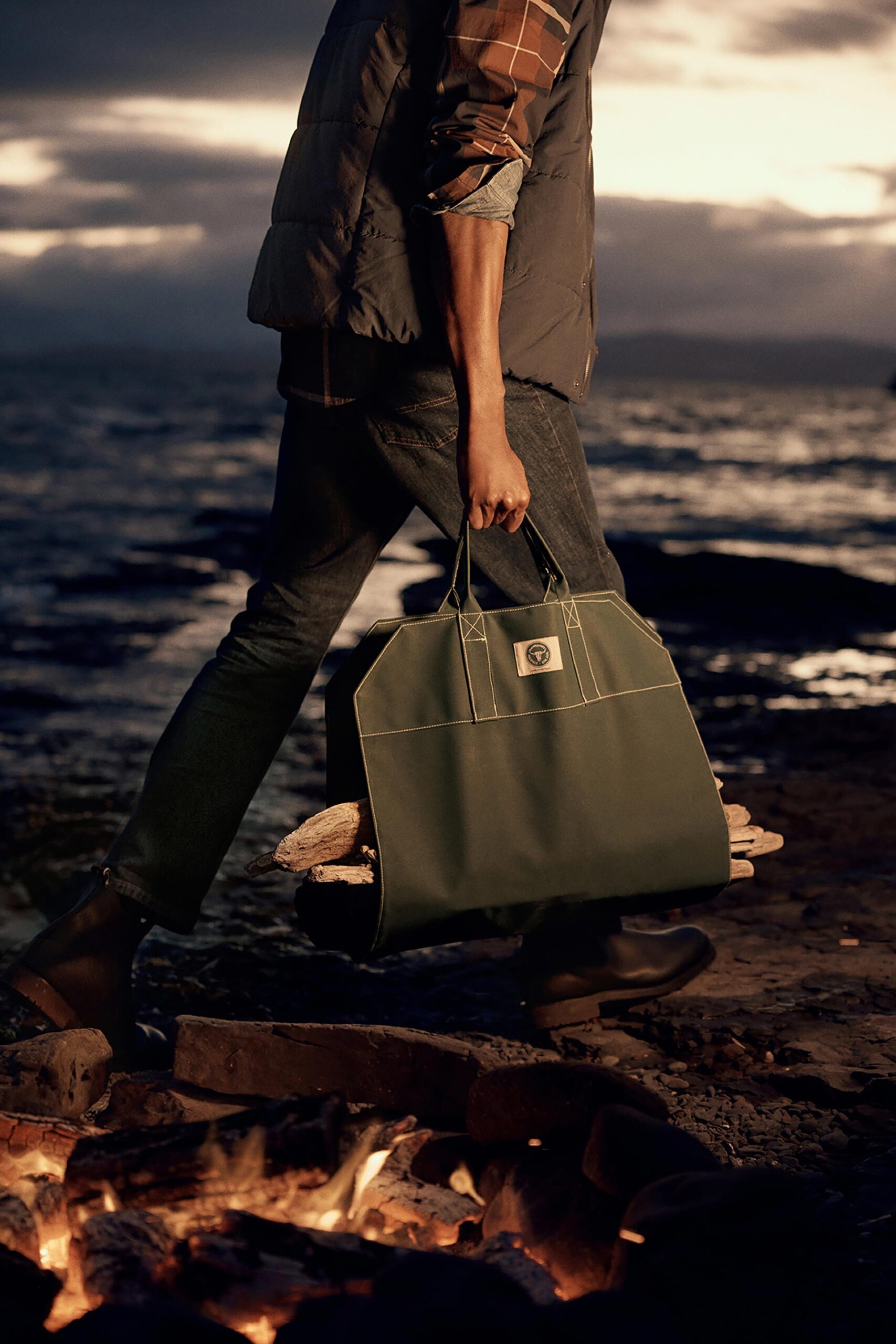 Man walks on rocky beach carrying firewood in olive drab tote bag near a bonfire at dusk. Casual style with puffer vest.