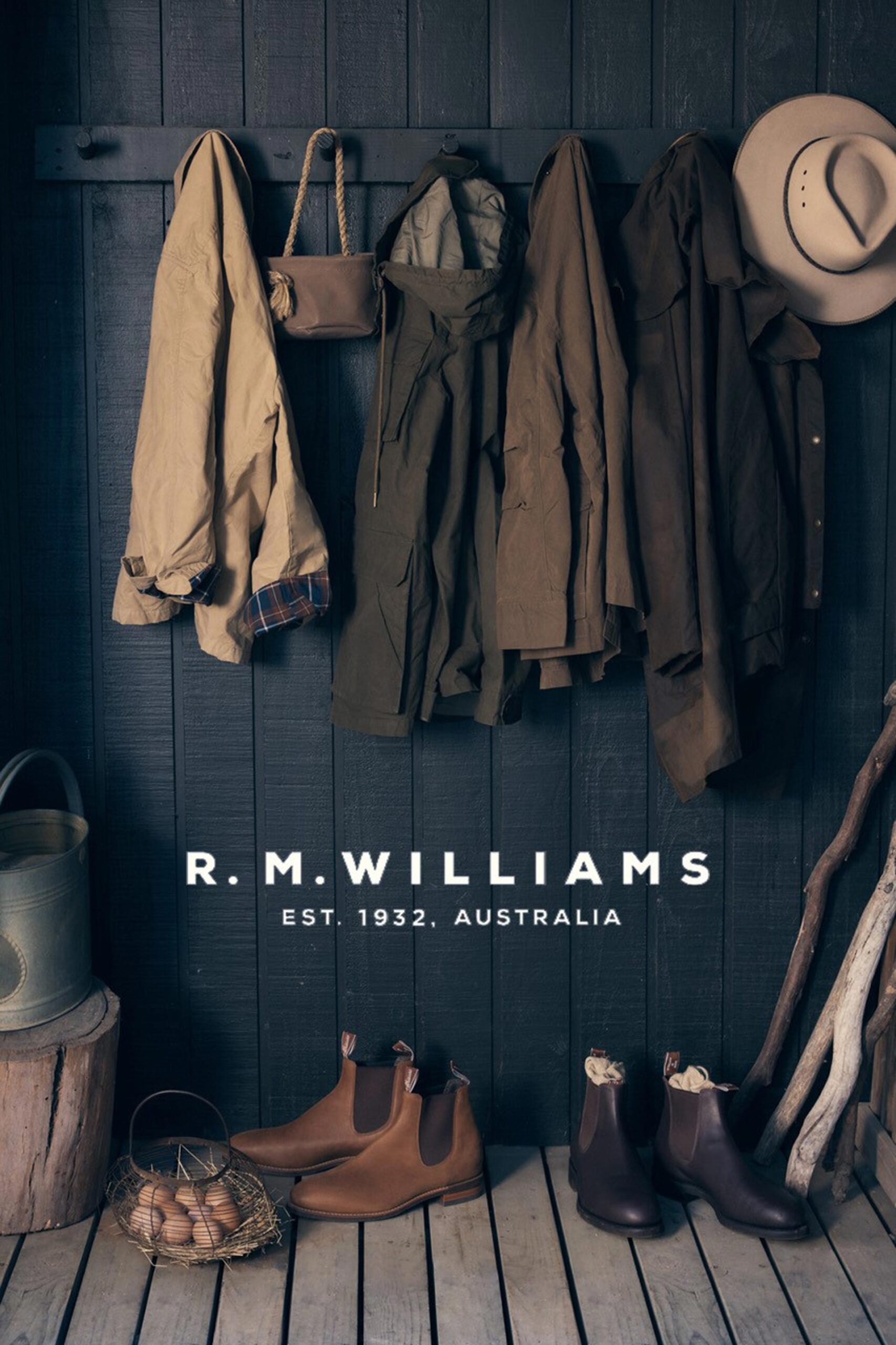Rustic product shot featuring R.M. Williams boots with hanging coats, a hat, and eggs in a basket, set against a dark wooden backdrop.