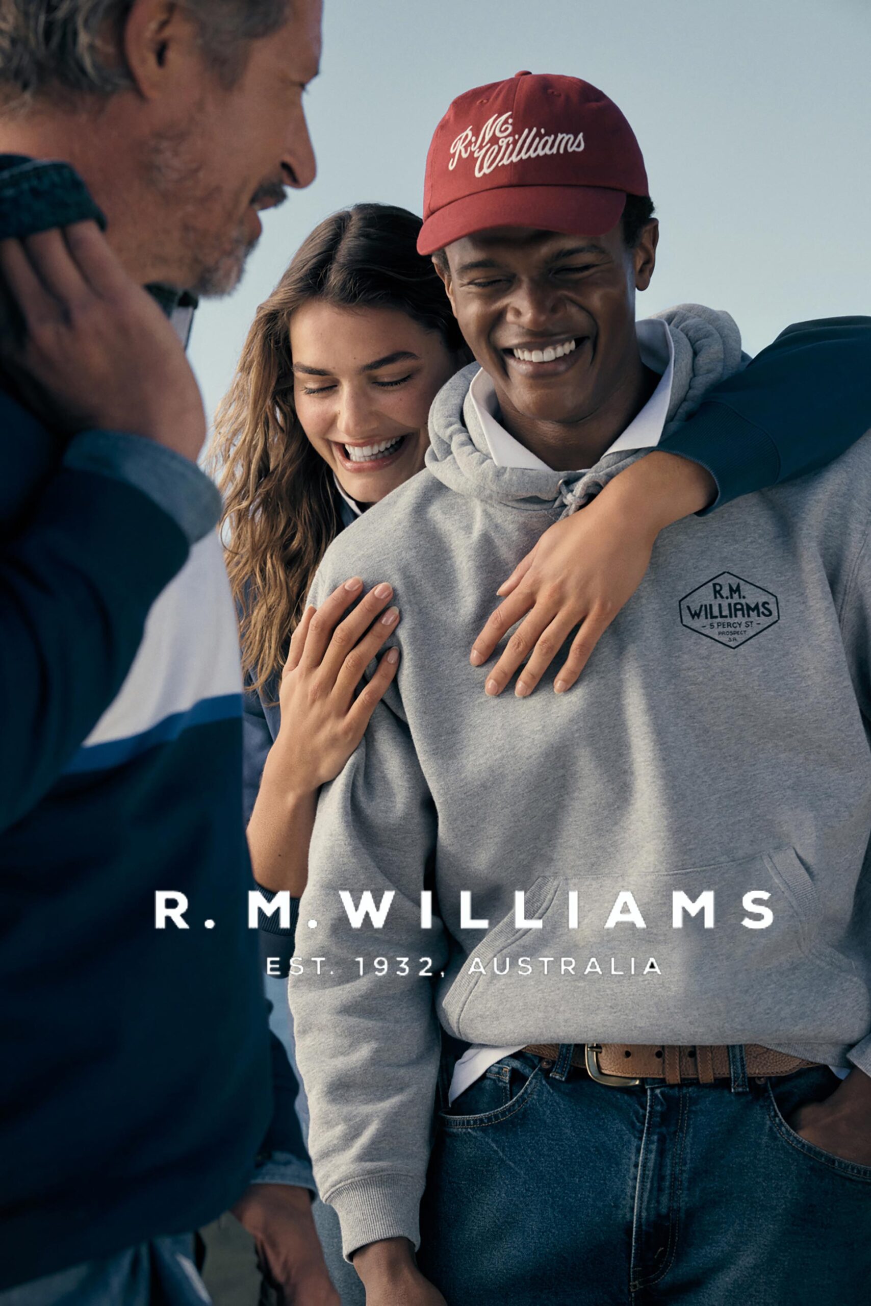 Three people, an adult man and woman flanking a smiling adult man outdoors, wearing a cap and grey hoodie, R.M. Williams brand visible.