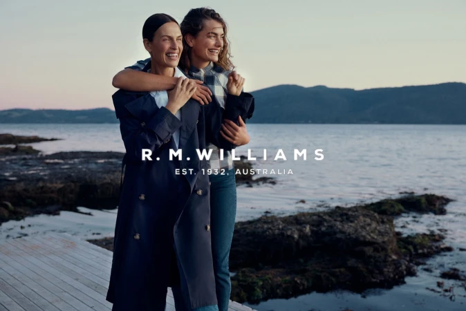 Two women stand embraced near a body of water at dusk, one in a blue trench coat, the other in a plaid shirt and jeans, smiling.