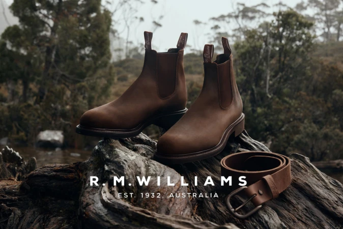 A pair of brown leather boots and a matching belt are displayed on a textured rock surface against an out-of-focus forest backdrop.