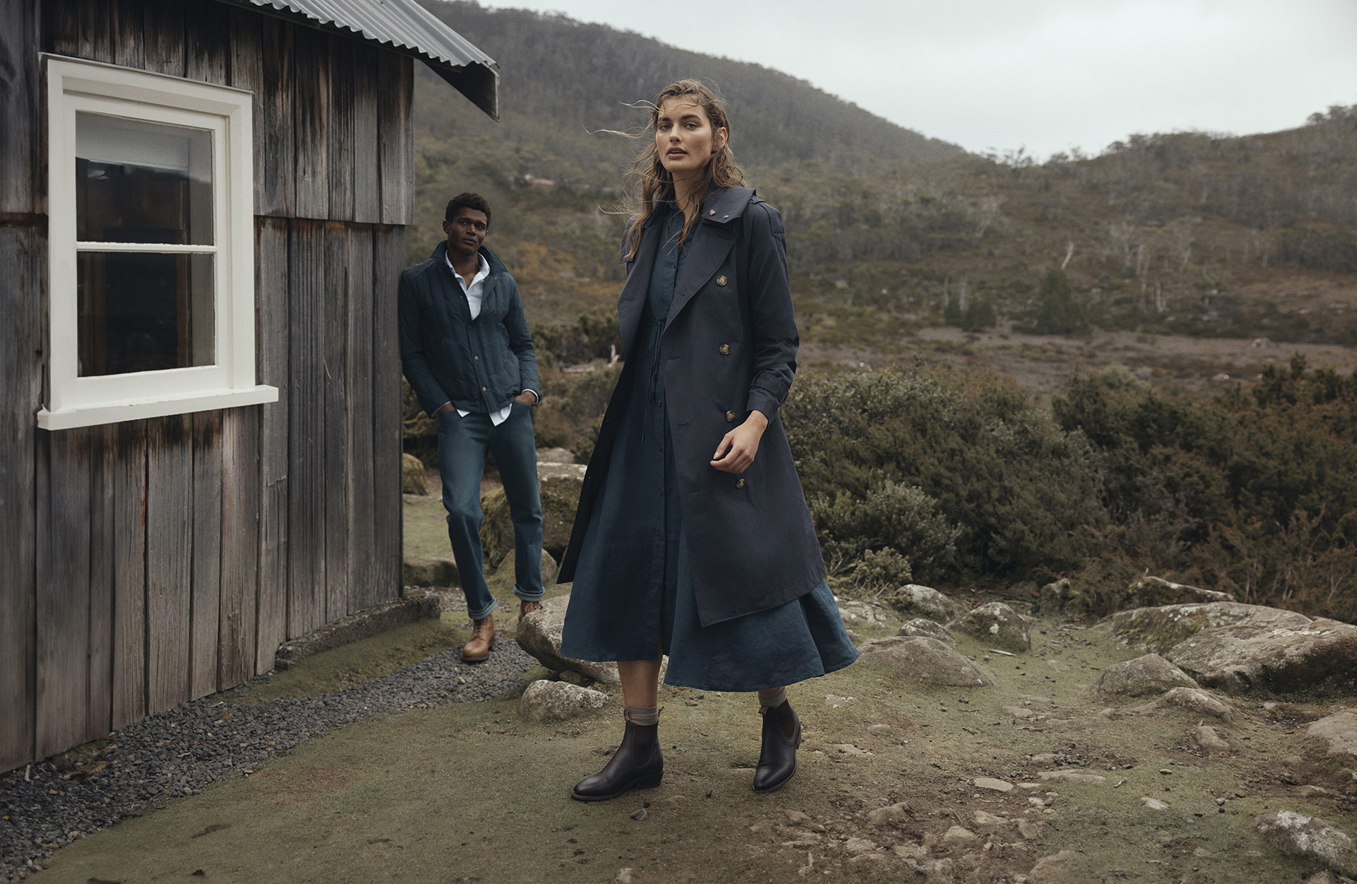 Fashion shot of a woman in a navy coat and dress standing outdoors with a man in jeans and a jacket next to a wooden structure.