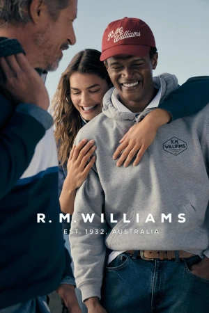 Black man smiling wearing a grey hoodie and red cap with a woman embracing him from behind and another person in the foreground.
