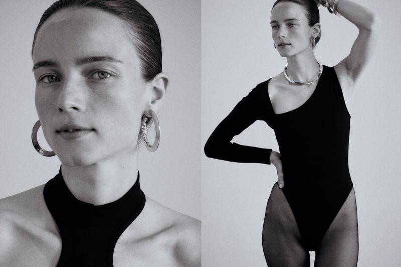 Black and white studio portrait of a woman wearing a black one-shoulder leotard, large hoop earrings, and bracelets against a neutral background.