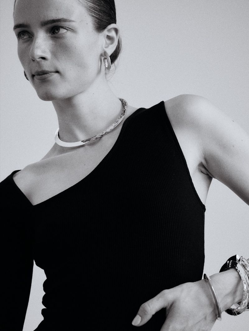 Monochrome portrait of a woman wearing a black, one-shoulder top with statement jewelry, including a collar necklace and bracelets.