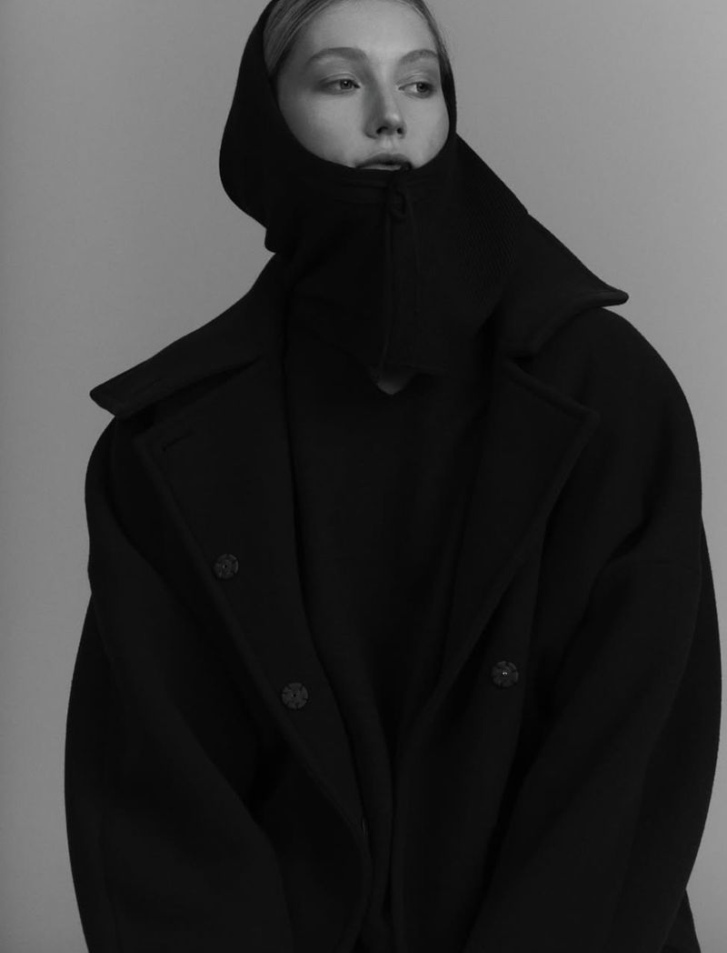 Monochrome portrait of a blonde woman wearing a dark overcoat with hood. Studio shot against a neutral gray backdrop, fashion photography.