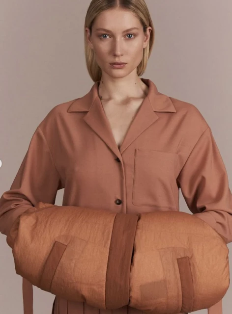 A blonde female poses indoors wearing a salmon shirt while holding a padded fabric object with bands, against a neutral backdrop.
