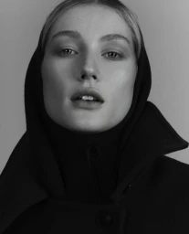 Close-up portrait of a woman wearing a dark hooded coat against a plain neutral background.