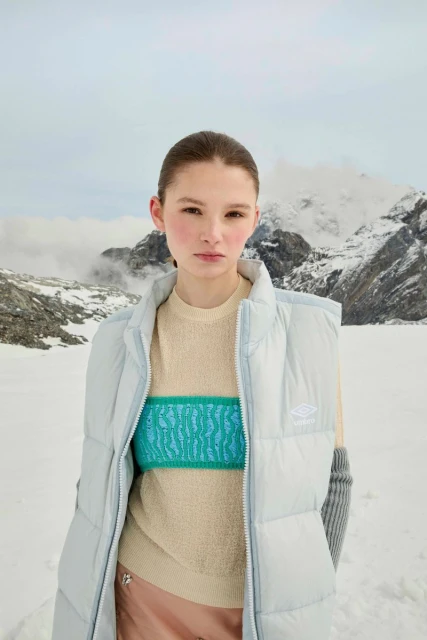A woman poses in a light blue puffer vest and knit sweater in a snowy mountain landscape with snow-covered peaks under a cloudy sky.