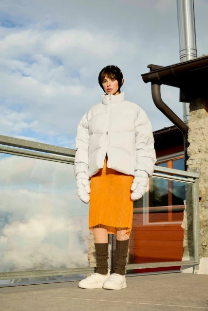 A brunette woman in a white puffer jacket, orange skirt, and white sneakers stands outside with a cloudy sky and mountain view in the background.