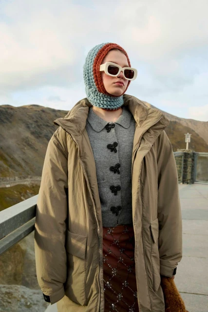 Woman in a crocheted balaclava and beige sunglasses wearing a beige trench coat standing in front of mountains on an overcast day.
