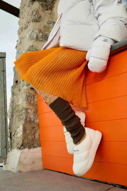 Fashionable person seated outdoors wearing a white puffer jacket, mustard skirt, and white puffy slippers with brown knitted leg warmers.