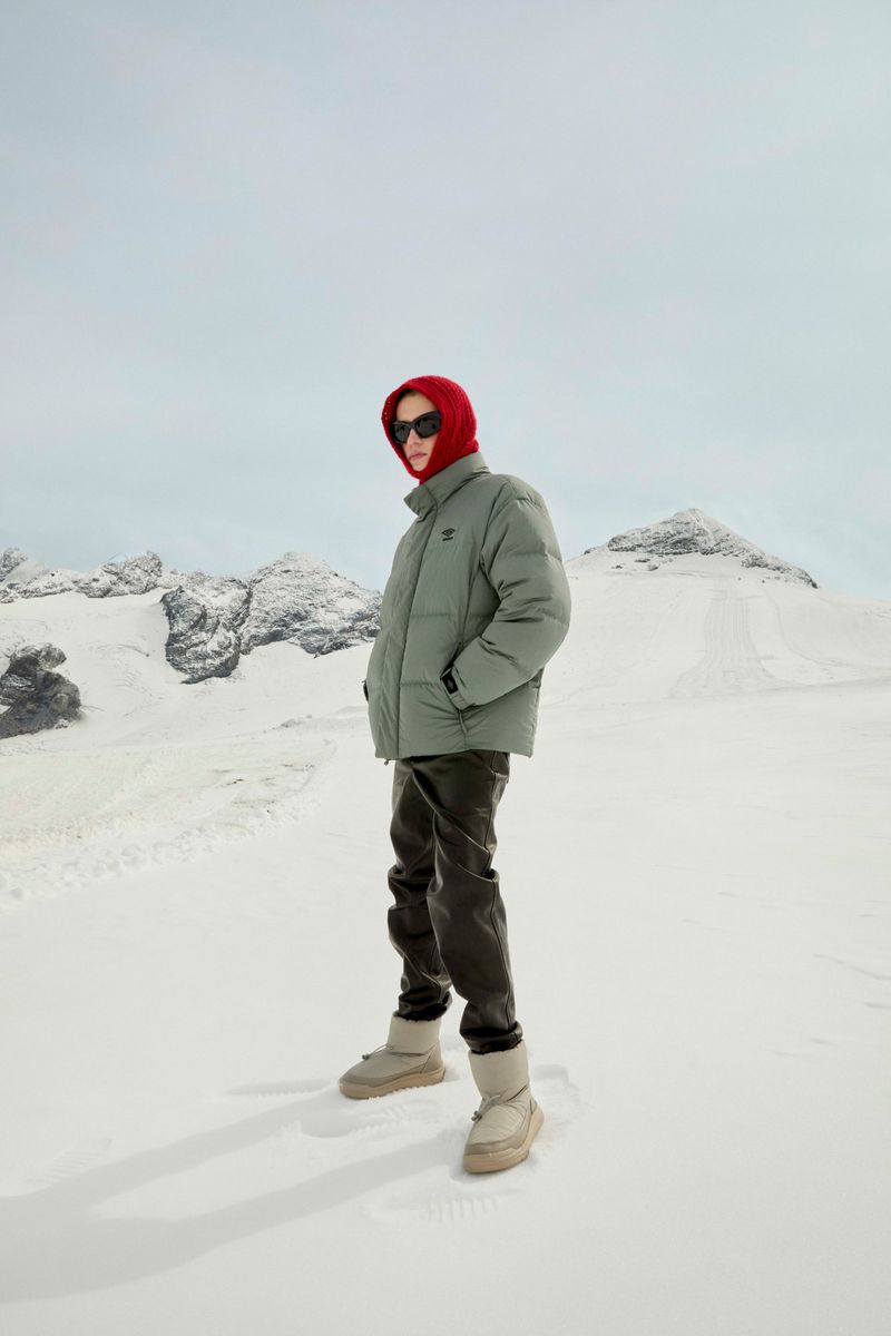 A person wearing a puffer jacket and boots stands in a snowy landscape with mountains, featuring winter apparel fashion outdoors.