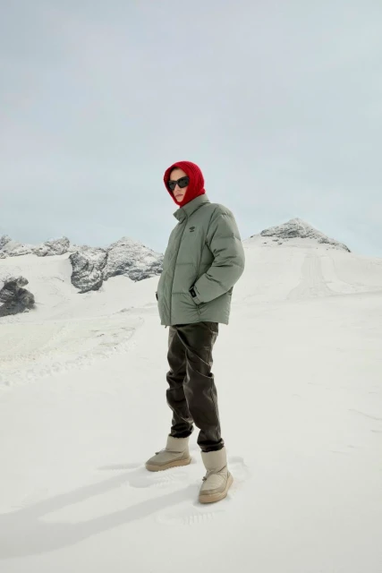 A person wearing a puffer jacket and boots stands in a snowy landscape with mountains, featuring winter apparel fashion outdoors.