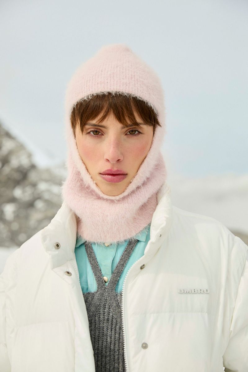 Close-up of a person wearing a light pink balaclava and a white puffer jacket, set against a blurry snow-covered mountain backdrop.