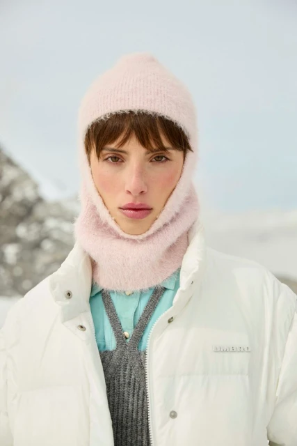 Close-up of a person wearing a light pink balaclava and a white puffer jacket, set against a blurry snow-covered mountain backdrop.
