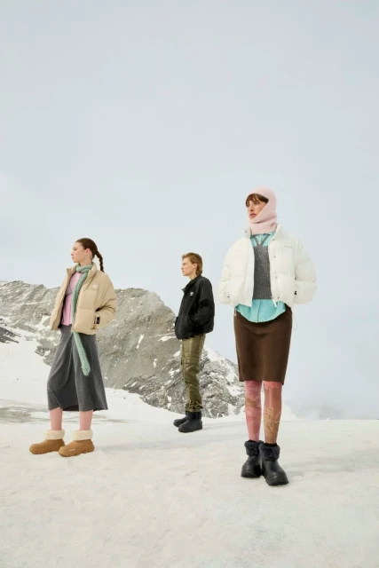 Three women model winter fashion in a snowy, mountainous landscape. They wear puffer jackets, skirts, and boots against a bright sky backdrop.