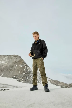 Male fashion model in a black sherpa jacket and olive cargo pants poses in a snowy, mountainous outdoor setting with sunglasses.