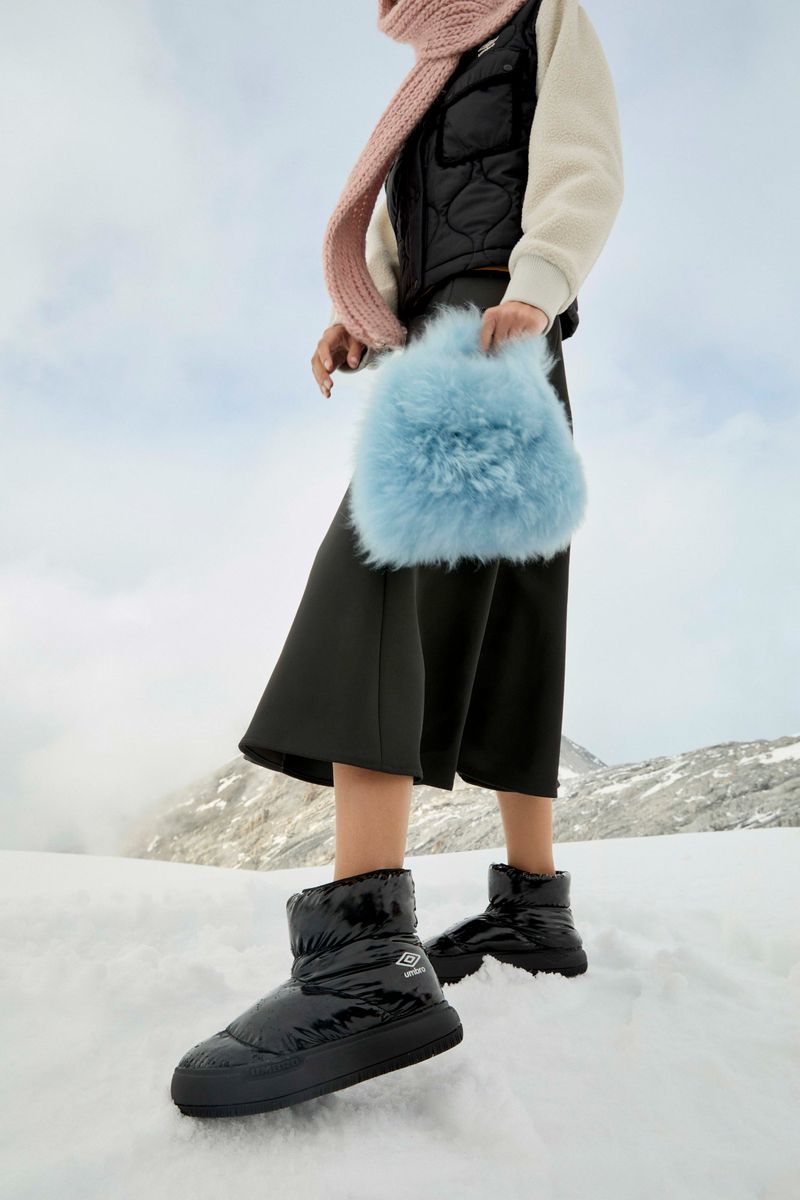 Woman in puffer boots, cropped pants, vest, and scarf stands in a snowy mountain landscape, holding a fuzzy light-blue handbag.