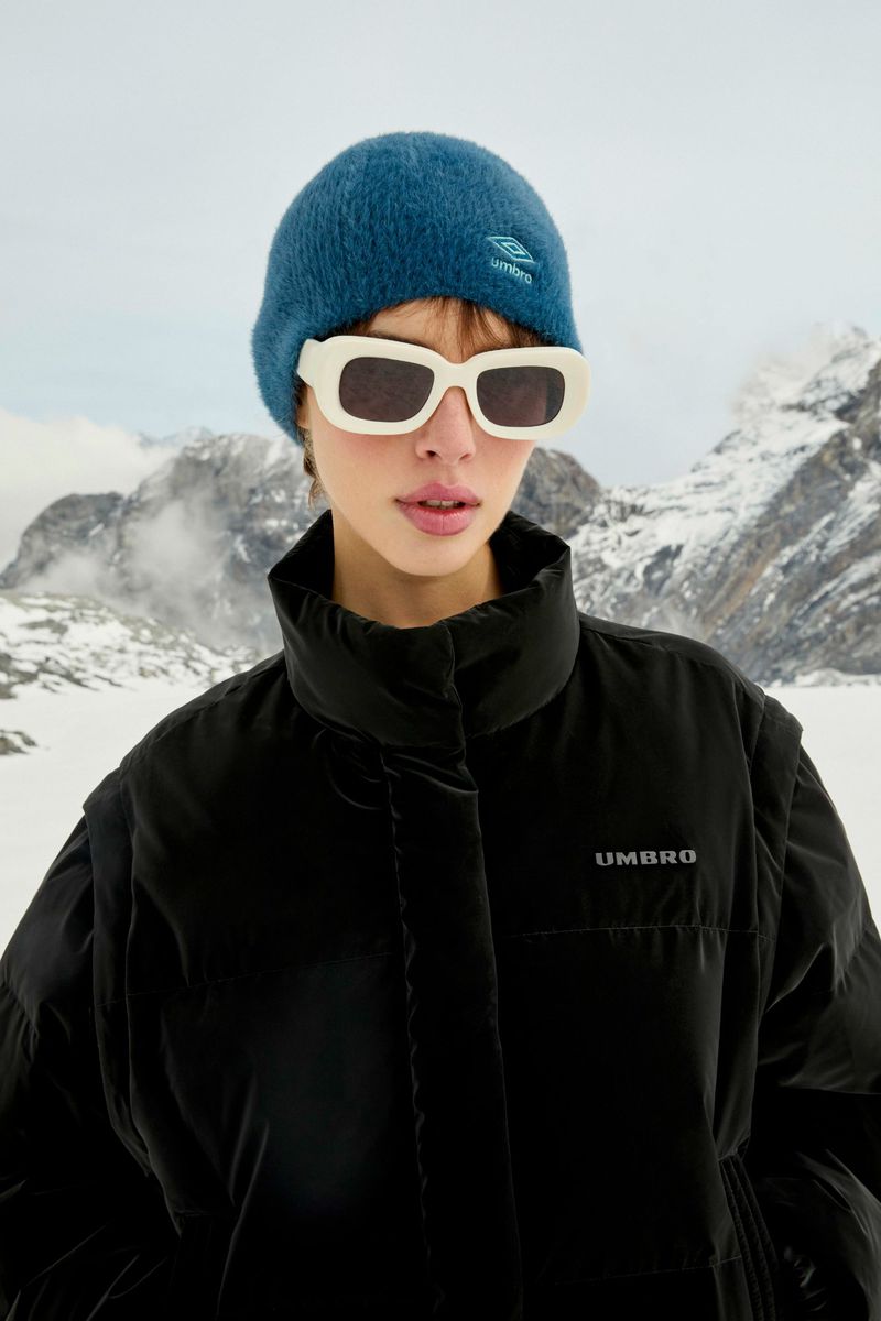 A woman models a black Umbro puffer jacket with mountains in the background, wearing sunglasses and a blue beanie. Outdoor fashion.