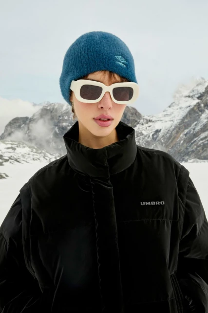A woman models a black Umbro puffer jacket with mountains in the background, wearing sunglasses and a blue beanie. Outdoor fashion.