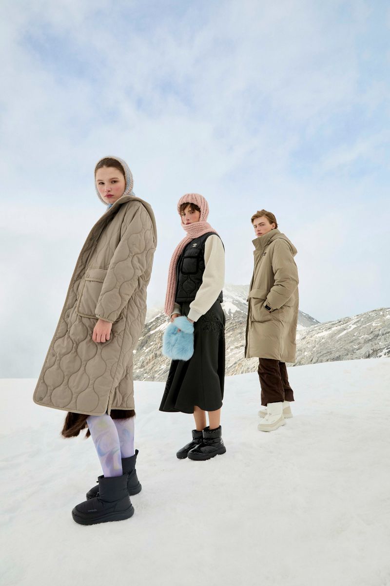 Three women in winter fashion pose in a snowy landscape, wearing coats, scarves, and boots against a backdrop of mountains and cloudy sky.