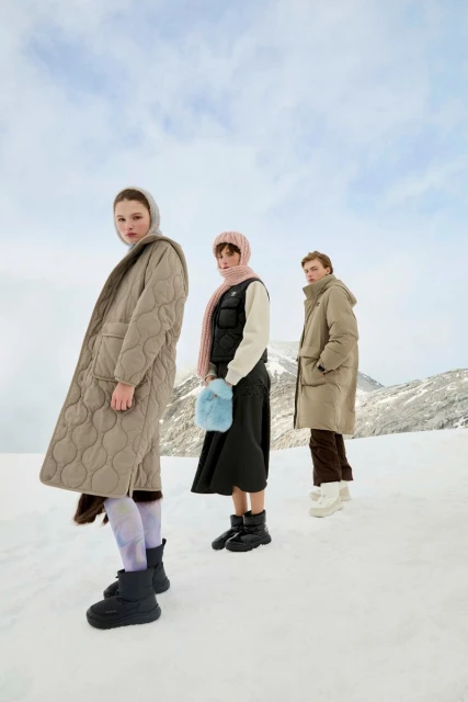 Three women in winter fashion pose in a snowy landscape, wearing coats, scarves, and boots against a backdrop of mountains and cloudy sky.