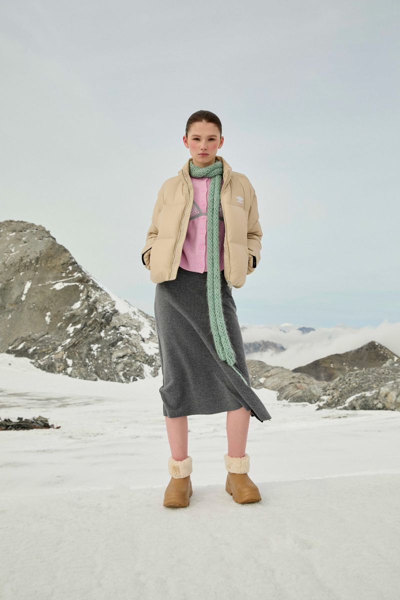 Young woman in a puffer jacket, skirt, and boots posing in a snowy, mountainous landscape. Light, neutral tones evoke a serene, outdoor fashion.