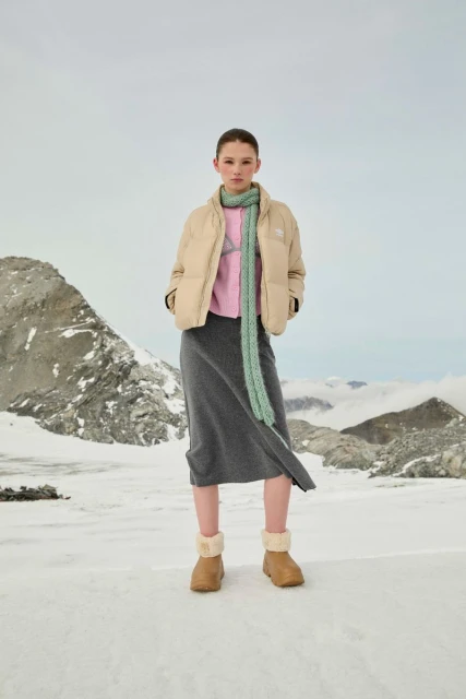 Young woman in a puffer jacket, skirt, and boots posing in a snowy, mountainous landscape. Light, neutral tones evoke a serene, outdoor fashion.