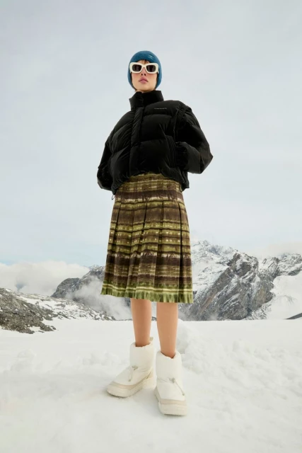 Fashionable woman poses in snow wearing a black puffer jacket, green pleated skirt, winter boots, white sunglasses, and a blue knit hat.