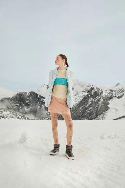 Fashion shot of a woman in a beige top, blue jacket, and light pink skirt posing outdoors in snowy mountains under a bright, clear sky.