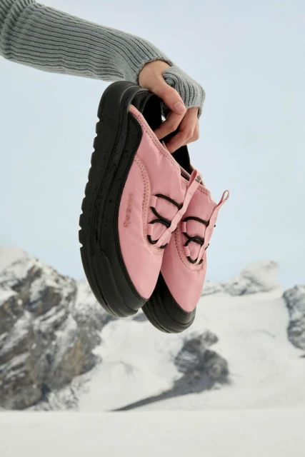 Close-up of light-pink athletic shoes with black soles being held up by a person wearing a grey long-sleeved shirt in front of snowy mountains.