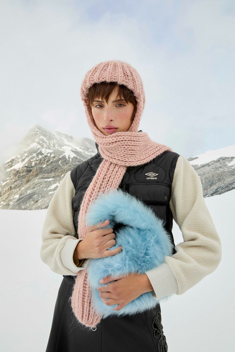 Woman in winter fashion poses in snow with mountains. She wears a pink knit hat and scarf, a black vest, and holds a blue furry purse.