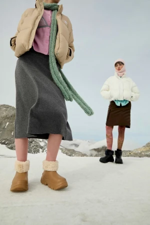 Two women pose in a snowy, mountainous environment, wearing winter coats, skirts, boots and hats. One stands closer to the camera.