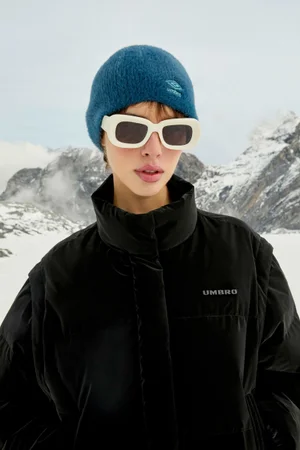 Model wearing a blue beanie, white sunglasses, and a black puffer jacket against a snowy mountain background.
