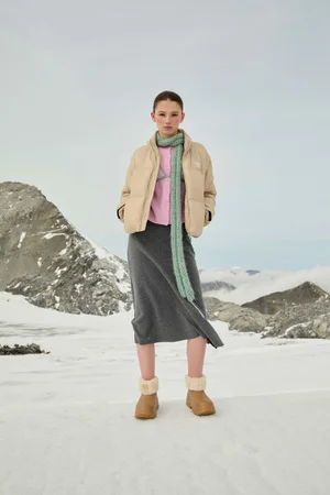 Model standing on a snowy mountain ridge wearing a beige puffer jacket, grey skirt, and winter boots against an overcast sky.