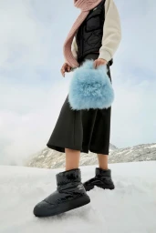 Model wearing black quilted puffer vest, dark skirt, and shiny black boots while holding a fluffy blue handbag in a snowy mountain landscape