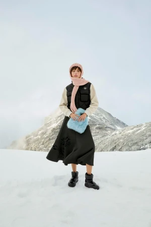 Model wearing a knit pink balaclava, black puffer vest, dark skirt, and holding a fluffy blue bag against a snowy mountain landscape