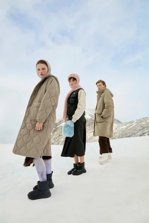 Three models wearing winter coats and padded outerwear standing on a snowy mountain landscape under a pale blue sky.