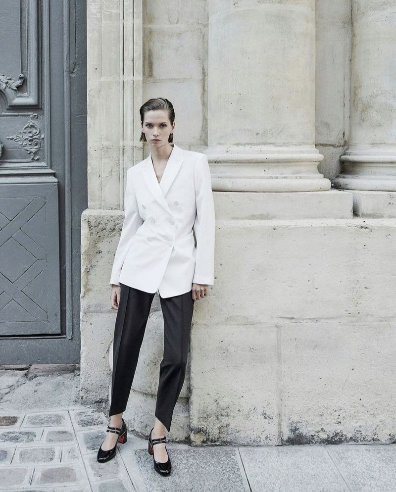 Fashion shot of a woman in a white blazer and dark pants posing in front of a stone building with columns and a blue door.