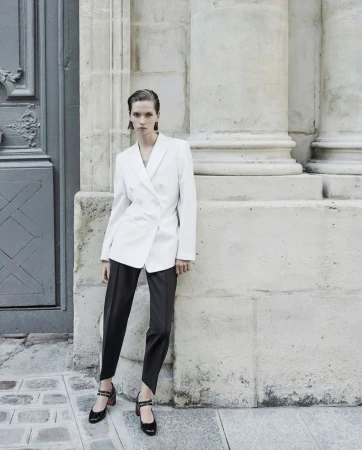 Fashion shot of a woman in a white blazer and dark pants posing in front of a stone building with columns and a blue door.