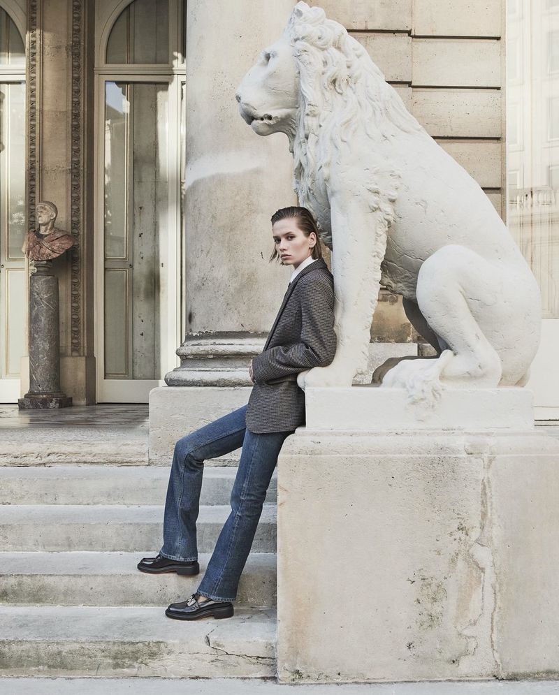 Fashion shot of a woman in a grey jacket and jeans sitting on stone steps next to a lion statue, in front of a building exterior.