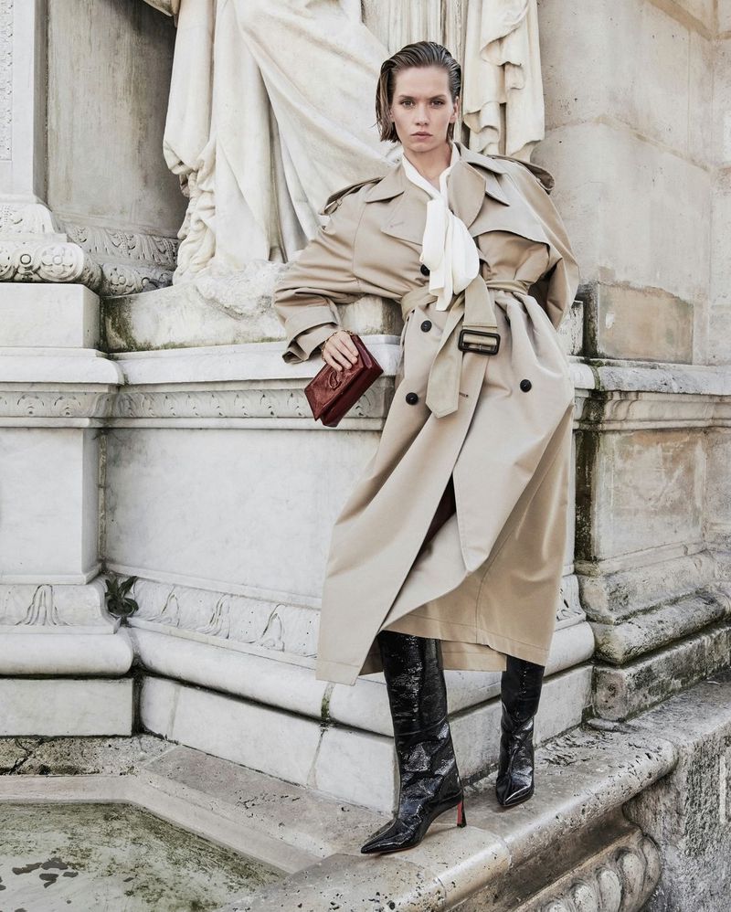 A woman in a beige trench coat holds a handbag while posing in front of a stone monument, featuring a statue, for a fashion shot.
