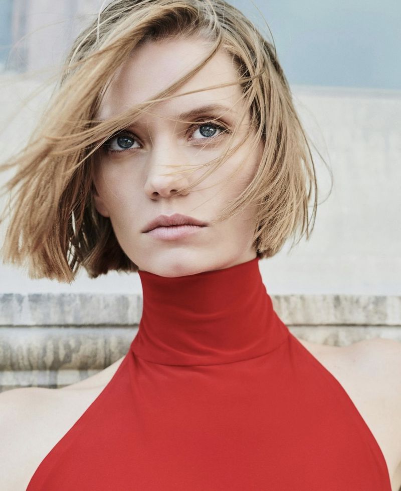 Close-up portrait of an adult woman with short blonde hair wearing a red high-necked dress. Background is a blurry outdoor setting.