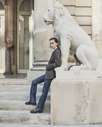 Model leaning against a large stone lion statue on outdoor steps, wearing a grey blazer and blue denim jeans.