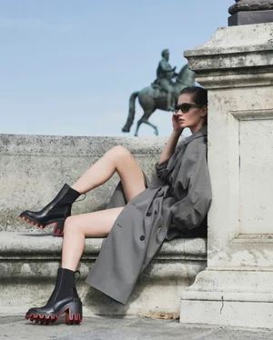 Model wearing dark sunglasses and black boots with red lug soles, seated against an outdoor stone monument.
