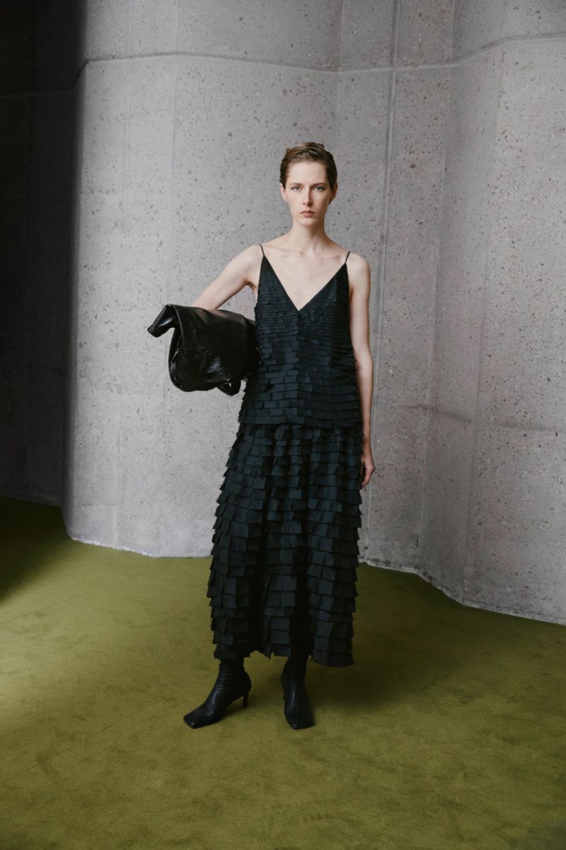 Woman in black tiered dress with black bag poses indoors against a textured wall and green floor; studio fashion shot.