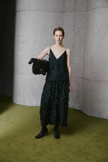 Woman in black tiered dress with black bag poses indoors against a textured wall and green floor; studio fashion shot.