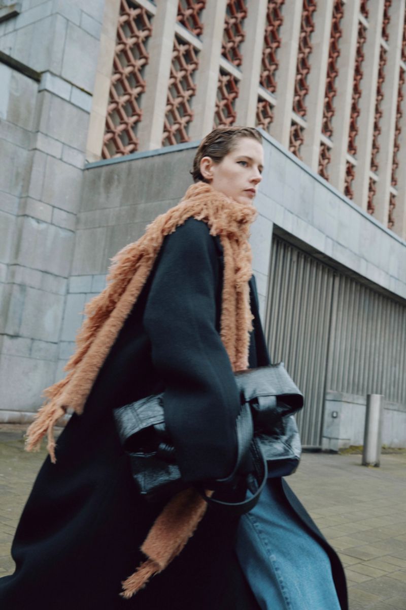 Woman in black coat with brown scarf and handbag walks on urban street with stone building in background during the daytime.